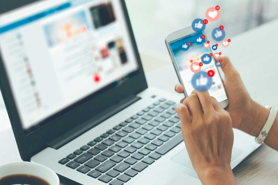 Person using a smartphone with social media notifications while a laptop displays a social media feed, representing the social media influence on Oman businesses.